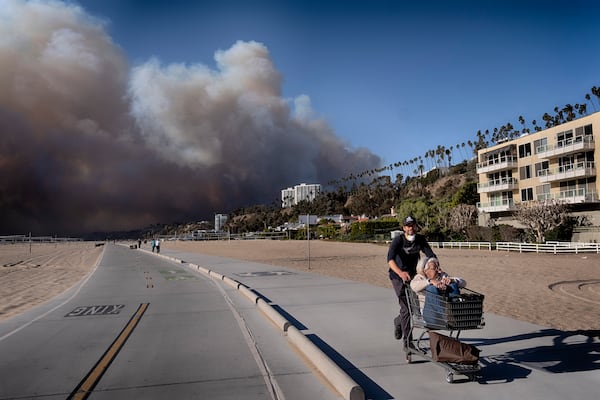 Jerome Krausse pushes his mother- in-law in a shopping cart as they evacuate from their home in the Pacific Palisades after a wildfire swept through their neighborhood in Santa Monica, Calif. on Tuesday, Jan. 7, 2025. (AP Photo/Richard Vogel)