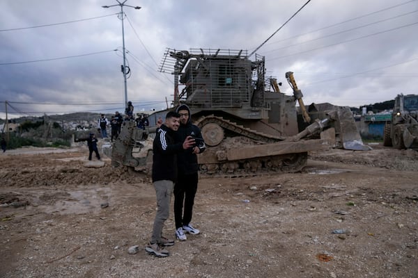 Two young Palestinian men pose for a photo with an Israeli bulldozer as it drives towards the occupied West Bank of Jenin camp, Sunday, Feb. 23, 2025. (AP Photo/Majdi Mohammed)