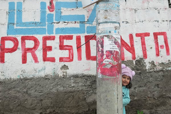 A girl hides behind a street post in front of a wall covered with election campaign signs for presidential candidate Luisa Gonzalez ahead of the presidential run-off election in Saquisili, Ecuador, Tuesday, Feb. 11, 2025. (AP Photo/Dolores Ochoa)