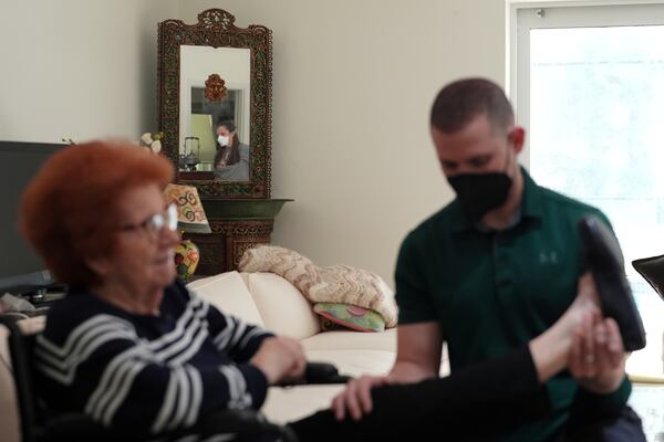 Kira Levin, 29, reflected in a mirror, wears a mask as her 98-year-old grandmother Jeanette Levin, left, does strength and endurance exercises with physical therapist assistant Alain Cabezas, in their home in Pinecrest, Fla., Monday, Dec. 16, 2024. (AP Photo/Rebecca Blackwell)