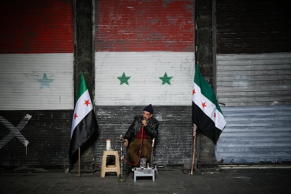 A man smokes a water pipe next to two Syrian "revolutionary" flags in front of a door still adorned with the official flag of the ousted government, near the Umayyad Mosque ahead of Friday prayers in Damascus, Syria, Friday, Dec. 20, 2024. (AP Photo/Leo Correa)