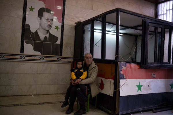 A man sits with a child next to a damaged image depicting the ousted Syrian President Bashar Assad, at the entrance of the Police headquarters, in Homs, Syria, Thursday, Dec. 26, 2024. (AP Photo/Leo Correa)