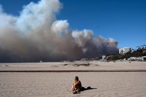 A lone sunbather sits and watches a large plume of smoke from a wildfire rise over the Pacific Palisades, in Santa Monica, Calif., on Tuesday, Jan. 7, 2025. (AP Photo/Richard Vogel)