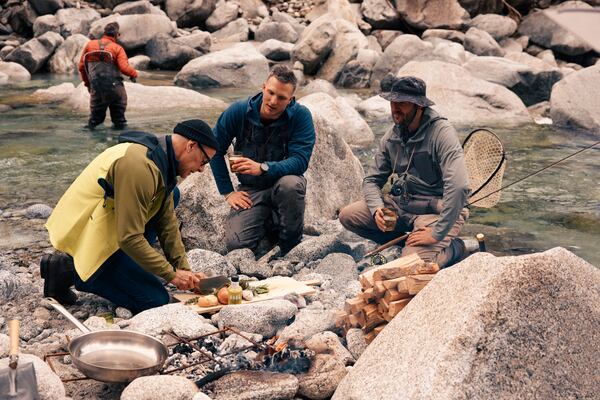 This image released by National Geographic shows Stanley Tucci, left, preparing a dish for anglers on the banks of the Sarca River during a visit to Italy's northernmost region, during the filming of his series "Tucci in Italy." (Matt Holyoak/National Geographic via AP)