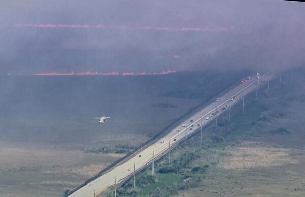 In this image taken from video provided by WSVN-TV, a brush fire burns along U.S. 1 as a helicopter flies above after dropping water Tuesday, March 18, 2025, south of Dade County, Fla. (WSVN-TV via AP)